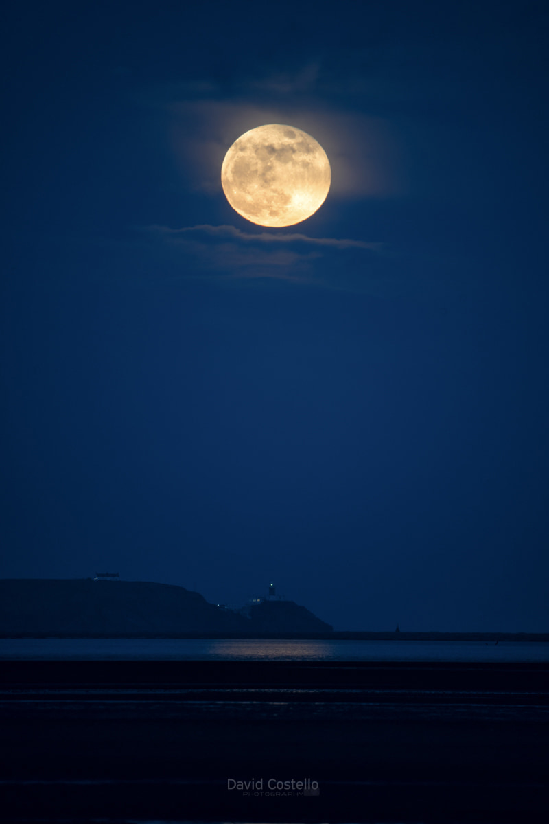 The full moon rising above the baily lighthouse viewed from across Dublin Bay in Sandymount.