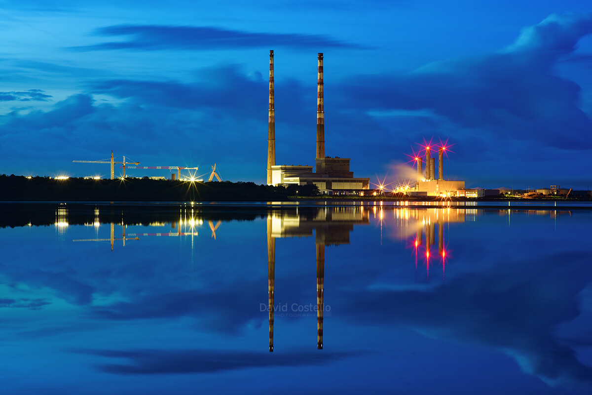 The Poolbeg Chimneys at twilight reflect in the tranquil waters along the strand as night falls and the lights come on.