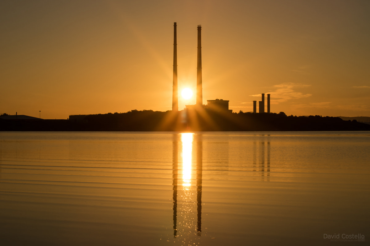 The summer sun rises between the iconic Poolbeg Chimneys and reflects in the water over Sandymount Strand and Dublin Bay.