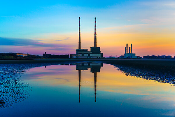 A dawn rainbow coloured sky above the Poolbeg Chimneys as the iconic towers reflect in the still water on Sandymount Strand.